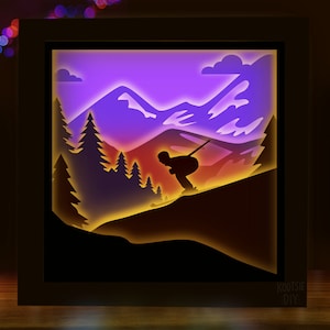 Könnte beinhalten: Ein gerahmter, beleuchteter Schattenkasten mit einem Skifahrer, der einen Berghang hinunterfährt. Die Szene ist als Silhouette vor einem Farbverlauf aus Lila, Orange und Gelb dargestellt, mit Kiefern und Berggipfeln. Der Rahmen ist dunkelbraun, und der Text "Kootie DIY" ist sichtbar.