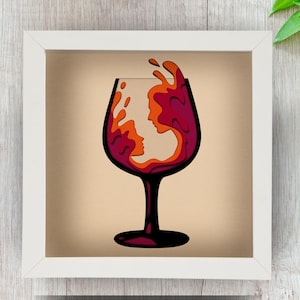 May include: A framed artwork featuring a layered paper design of a wine glass. The glass is filled with red and orange shapes, suggesting wine and a face profile. The artwork is set against a beige background.