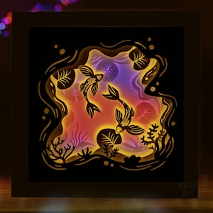 May include: A black wooden frame with a 3D paper cut-out design of two koi fish swimming in a pond. The fish are silhouetted against a colorful background of pink, purple, and yellow.