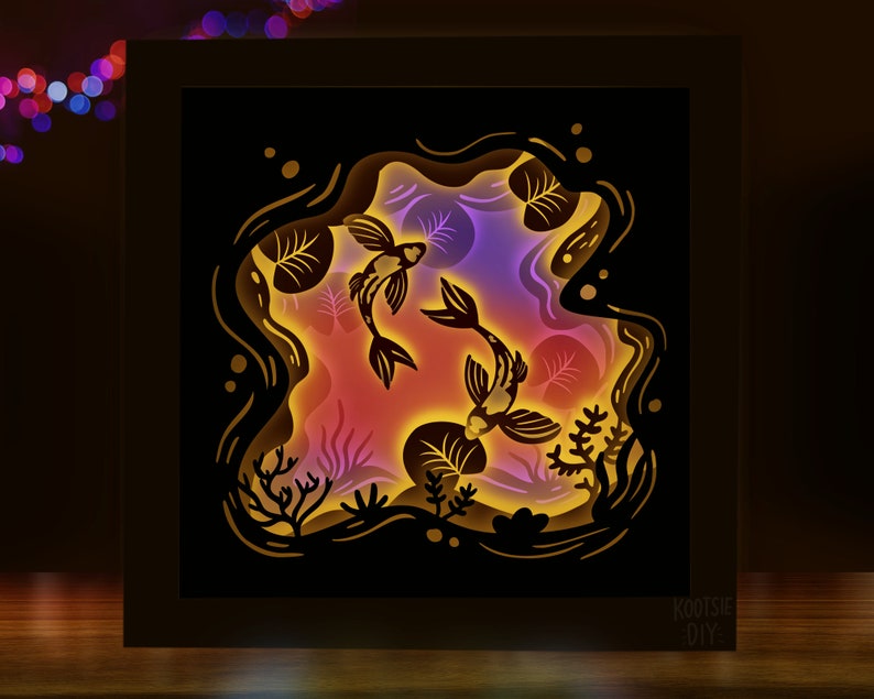 May include: A black wooden frame with a 3D paper cut-out design of two koi fish swimming in a pond. The fish are silhouetted against a colorful background of pink, purple, and yellow.