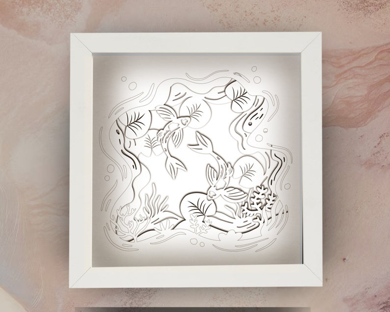 May include: White frame with a white background and a black paper cut-out design of two fish swimming in a pond with lily pads and water plants.