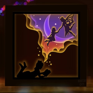 May include: A 3D layered papercraft nightlight featuring a girl reading a book and a fairy flying with another girl on a crescent moon. The nightlight is illuminated with warm yellow light and has a dark brown wooden frame. The text "KOOTSIE DIY" is visible in the bottom right corner.