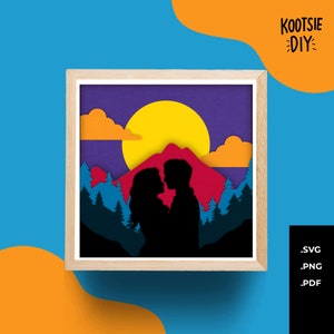 Op de afbeelding: Een ingelijst papieren kunstwerk met een silhouet van een stel dat elkaar omhelst voor een bergketen, een gele zon en oranje wolken. De achtergrond is een verloop van paars en blauw. Het frame is van licht hout. De tekst "KOOTSIE DIY" staat rechtsboven.