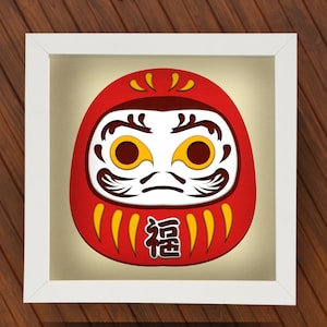 May include: A white framed picture of a red and yellow daruma doll with the Japanese character for "good fortune" written on it.