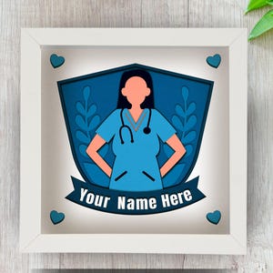 Puede incluir: Una obra de arte enmarcada que representa a una enfermera con uniforme azul y estetoscopio. El diseño incluye una forma de escudo con elementos florales y una pancarta que dice "Your Name Here". El marco es blanco y la obra tiene un efecto 3D.