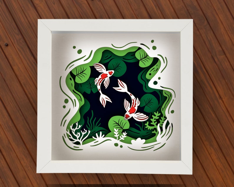 May include: A white frame with a 3D paper cut-out design of two white and red fish swimming in a green pond with lily pads. The pond is surrounded by green and white foliage.