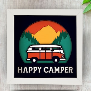 May include: A framed art piece featuring a retro orange and white camper van set against a backdrop of mountains and trees. The words "HAPPY CAMPER" are displayed in white at the bottom. The artwork is layered with a white frame.