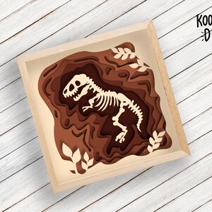 Pode incluir: Um esqueleto de dinossauro de papel castanho num quadro de papel em camadas. O quadro &eacute; feito de papel castanho e tem um fundo branco. O esqueleto de dinossauro est&aacute; em p&eacute; com a cabe&ccedil;a inclinada para o lado. O quadro tem o texto "KOOTSIE DIY" no canto superior direito.