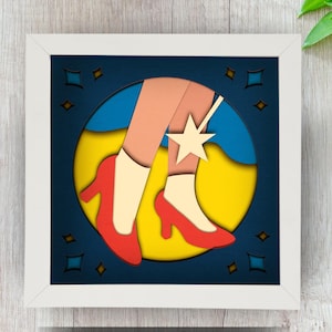 May include: A white frame with a blue background and a yellow, red, and white paper cutout of a pair of red shoes with a star on the ankle.