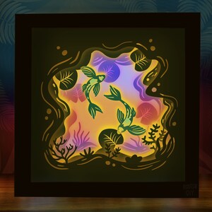 May include: A 3D paper cut-out art piece depicting two green fish swimming in a pond with lily pads and other aquatic plants. The art piece is illuminated from behind, creating a glowing effect.