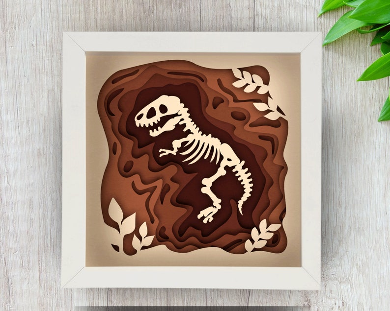 Pode incluir: Uma moldura branca com um recorte de papel castanho e bege de um esqueleto de dinossauro no interior. O dinossauro est&aacute; numa pose como se estivesse a correr. A moldura est&aacute; sobre uma superf&iacute;cie de madeira clara.