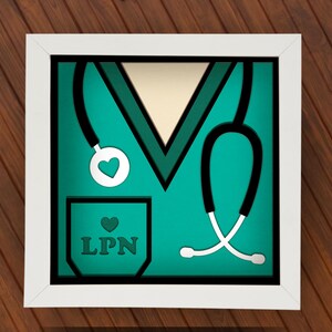 May include: A framed teal and white artwork featuring a stylized medical scrub top design. The artwork includes a stethoscope, a pocket with the letters "LPN", and heart-shaped accents. The frame is white, and the background is a wooden surface.