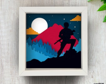 May include: A framed paper art piece depicting a silhouette of a person with a rifle on a mountain. The background shows a red mountain, a full moon, clouds, and a forest. The artwork is presented in a white frame.