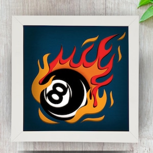 May include: A white frame with a blue background and a black 8-ball with flames around it. The flames are red, orange, and yellow.