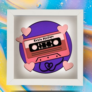 May include: A framed, layered paper art piece featuring a pink cassette tape design with the text "Kayla Mixtape" on a purple background. Pink hearts surround the cassette, with a heart-shaped tape detail.