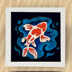 Pode incluir: Obra de arte em papel emoldurada com um design de carpa koi. O peixe é branco com marcas vermelhas, contra um fundo de água azul. A obra de arte está em uma moldura quadrada branca, criando um efeito 3D. Medidas: aproximadamente 20 cm x 20 cm.