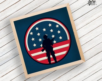 May include: A framed paper craft featuring a silhouette of a soldier standing in front of a red, white and blue American flag. The flag is in a circle shape with white stars on a blue background.