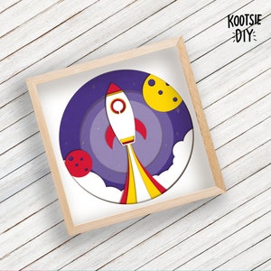 May include: A paper craft rocket ship with yellow and red flames, flying through a purple and blue space scene with a yellow moon and a red planet. The scene is framed in a light wood frame. The text "KOOTSIE DIY" is in the upper right corner of the image.
