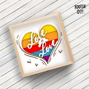 May include: A white paper heart with a rainbow layered design and the words "Love is Love" cut out. The heart is framed in a light wood frame.
