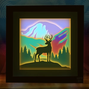 May include: A framed, layered paper art piece featuring a silhouette of a deer with large antlers standing on a hill, with a mountain and forest backdrop. The artwork uses shades of green, blue, purple, and yellow. The text "KOOTSIE DIY" is in the bottom right corner.