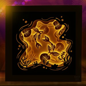 May include: A black framed 3D paper art piece depicting two gold fish swimming in a pond with lily pads. The fish are cut out and layered to create a 3D effect. The background is a dark blue with white stars.