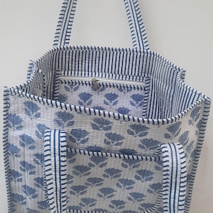 以下が含まれることがあります： ストライプのハンドルとトリムが付いた、青と白の花柄のトートバッグ。バッグには、ジッパー付きの内ポケットと外側の花柄模様があります。バッグはキルティング生地で作られています。