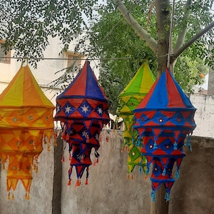 May include: Four colourful fabric lanterns with geometric patterns and tassels hanging from a tree branch. The lanterns are in shades of yellow, red, blue, and green.