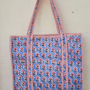 K&ouml;nnte beinhalten: Eine blaue und rosafarbene Blumentasche mit rot-wei&szlig; gestreiften Henkeln. Die Tasche ist aus Baumwolle und hat ein gestepptes Design.
