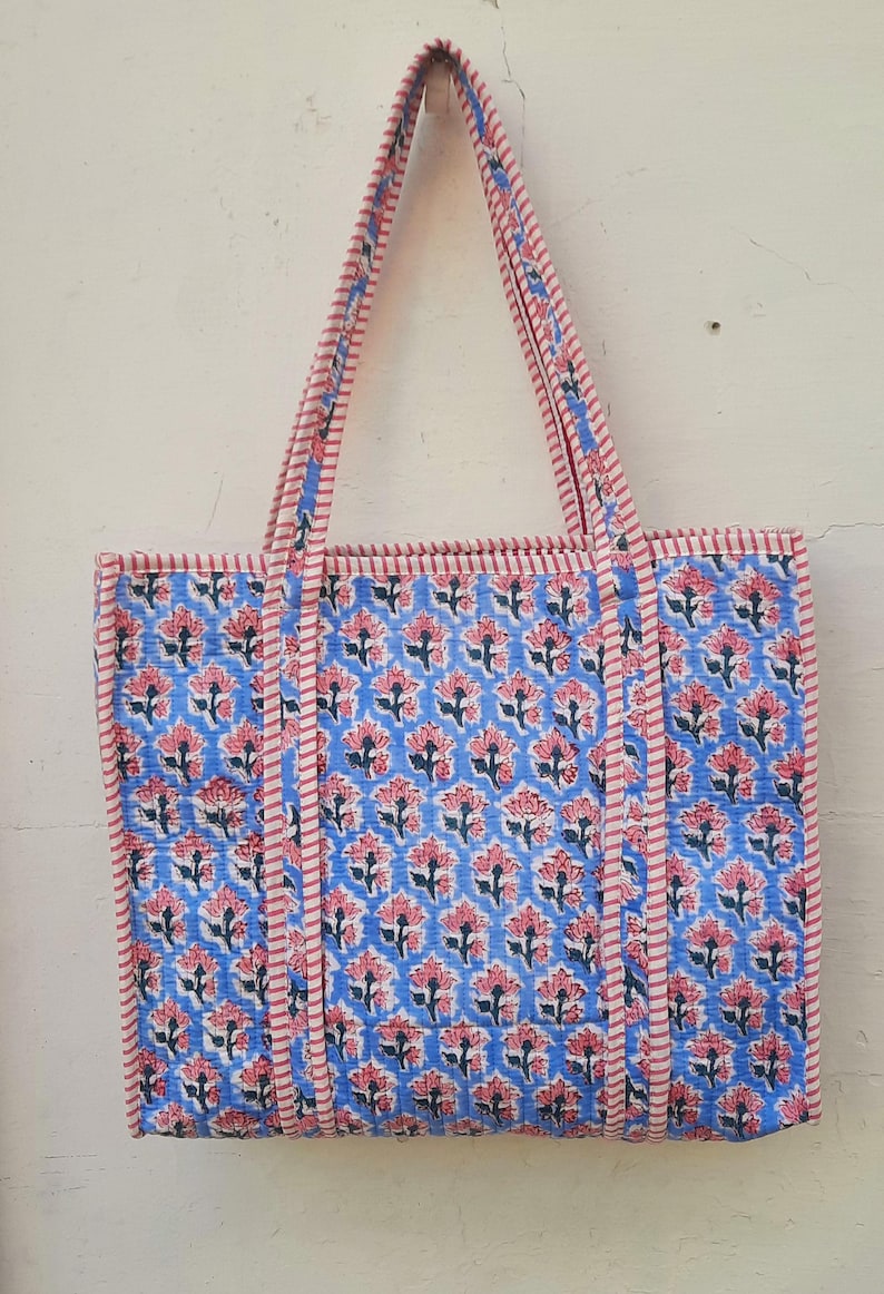 K&ouml;nnte beinhalten: Eine blaue und rosafarbene Blumentasche mit rot-wei&szlig; gestreiften Henkeln. Die Tasche ist aus Baumwolle und hat ein gestepptes Design.