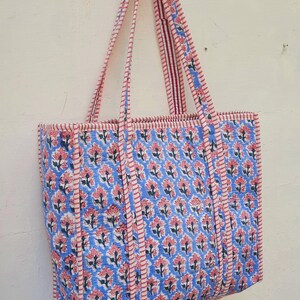 K&ouml;nnte beinhalten: Eine blaue und rosafarbene Blumentasche mit rot-wei&szlig; gestreiften Henkeln. Die Tasche ist aus Baumwolle und hat ein gestepptes Design.