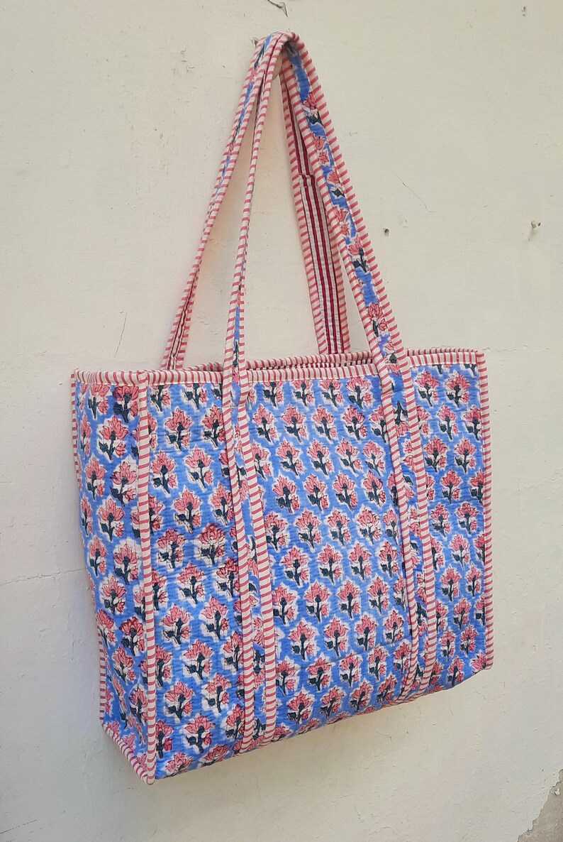 K&ouml;nnte beinhalten: Eine blaue und rosafarbene Blumentasche mit rot-wei&szlig; gestreiften Henkeln. Die Tasche ist aus Baumwolle und hat ein gestepptes Design.