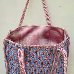 K&ouml;nnte beinhalten: Eine blaue und rosafarbene Tragetasche mit floralem Muster und rot-wei&szlig; gestreiften Henkeln. Die Tasche ist aus Baumwolle und hat ein gestepptes Design.