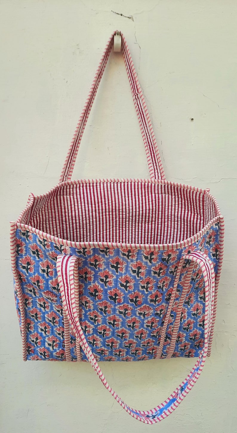 K&ouml;nnte beinhalten: Eine blaue und rosafarbene Tragetasche mit floralem Muster und rot-wei&szlig; gestreiften Henkeln. Die Tasche ist aus Baumwolle und hat ein gestepptes Design.