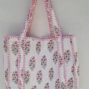 May include: A white cotton tote bag with pink and white striped trim. The bag features a floral print with pink and green flowers.
