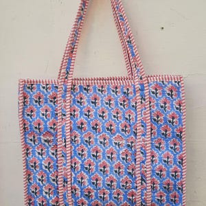 May include: A blue and pink floral tote bag with white stripes on the straps. The bag is made of cotton and has a quilted design.
