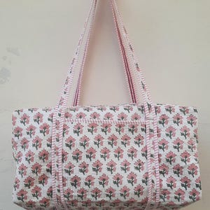 Może przedstawiać: Biała bawełniana torba typu tote z nadrukiem różowych i zielonych kwiatów. Torba ma dwa różowe i białe paski.