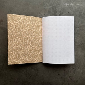 May include: An open notebook with a brown cover featuring a white floral pattern. The inside pages are white with a dotted grid pattern.