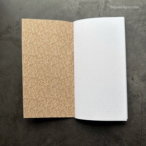 Puede incluir: Un cuaderno abierto con una cubierta marrón con un patrón floral blanco. Las páginas interiores son blancas con puntos grises espaciados uniformemente.