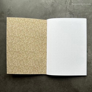 Puede incluir: Un cuaderno abierto con una cubierta marrón con un patrón floral blanco. La página opuesta es blanca con puntos grises espaciados uniformemente.