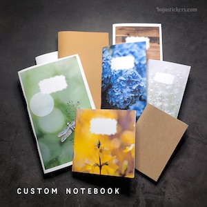 Op de afbeelding: Een set van zes gepersonaliseerde notitieboekjes met verschillende ontwerpen. De notitieboekjes hebben een bruine papieren omslag, een groene omslag met een libel, een blauwe omslag met hortensia's, een gele omslag met een bloem, een witte omslag met witte bloemen en een bruine omslag met een houtnerfdesign. De notitieboekjes hebben een blanco label aan de voorkant voor personalisatie.  CUSTOM NOTEBOOK is onderaan de afbeelding gedrukt.