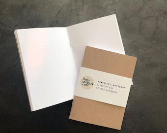 May include: An open, dot-grid notebook next to a closed, brown Traveler's Notebook. The closed notebook has a white band with the text "bujostickers.com" and "TRAVELER'S NOTEBOOK PASSPORT SIZE DOTTED NUMBERED". The open notebook has dotted pages.