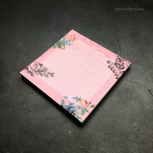 Könnte beinhalten: Rosa florale Haftnotizen mit einem weißen Zentrum zum Schreiben. Das Blumenmuster zeigt rosa, blaue und graue Blumen.