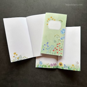 以下が含まれることがあります： 花柄の表紙が付いた空白のノートが3冊あります。表紙は緑色で、ブルーベル、勿忘草、赤いベリーなど、さまざまな花が描かれています。ノートは開いており、中の空白のページを見ることができます。