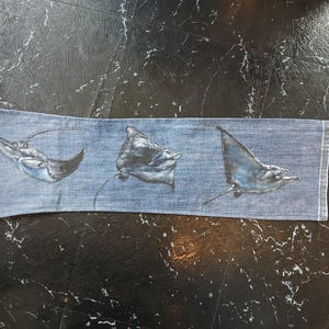 May include: A rectangular piece of light blue denim fabric featuring three hand-painted manta rays in shades of dark blue, grey, and white. The rays are depicted in various swimming positions across the horizontal length of the denim material.
