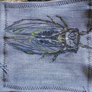 May include: A denim patch with a hand-painted insect design. The insect is depicted in shades of blue, green, and yellow. The square patch has a zigzag stitched border.