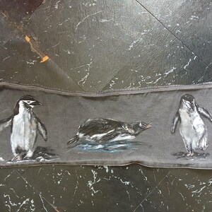 May include: A rectangular piece of dark grey fabric featuring a hand-painted illustration of three penguins. One penguin stands on the left, one swims in the centre, and one stands on the right. The fabric has visible stitching along the edges.