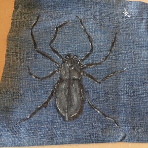 May include: A piece of blue denim fabric featuring a hand-painted black spider illustration. The spider is depicted with a central body and eight long, thin legs extending outward. The paint has a slightly glossy finish against the textured fabric.