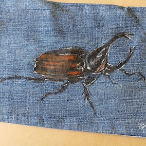 May include: A denim fabric with a hand-painted rhinoceros beetle design. The beetle is painted in brown, black, and white, with detailed legs and antennae. The artwork is centered on the blue denim.