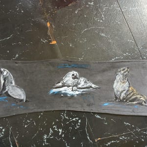 May include: A rectangular dark grey fabric panel featuring a hand-painted illustration of three seals. The left seal is upright, the centre seal is lying down, and the right seal is looking upward. Each seal is accented with small blue paint strokes.