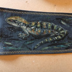 May include: A rectangular, dark blue fabric item featuring a detailed painting of a lizard. The lizard has a brown and green pattern. The edges are stitched with gold thread. The lizard is painted on a dark blue background.